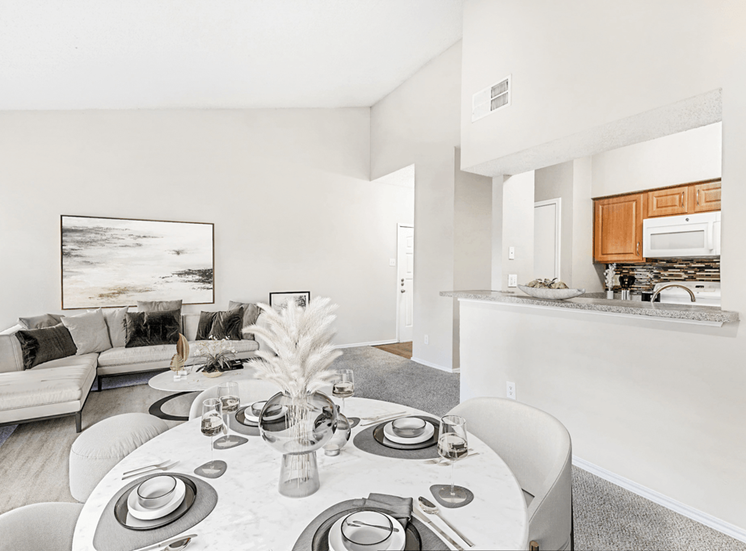 Virtually staged dining and livingroom with carpet, white table with chairs, sectional couch, granite style countertop on breakfast bar, light wood cabinetry and white appliances with vaulted ceiling